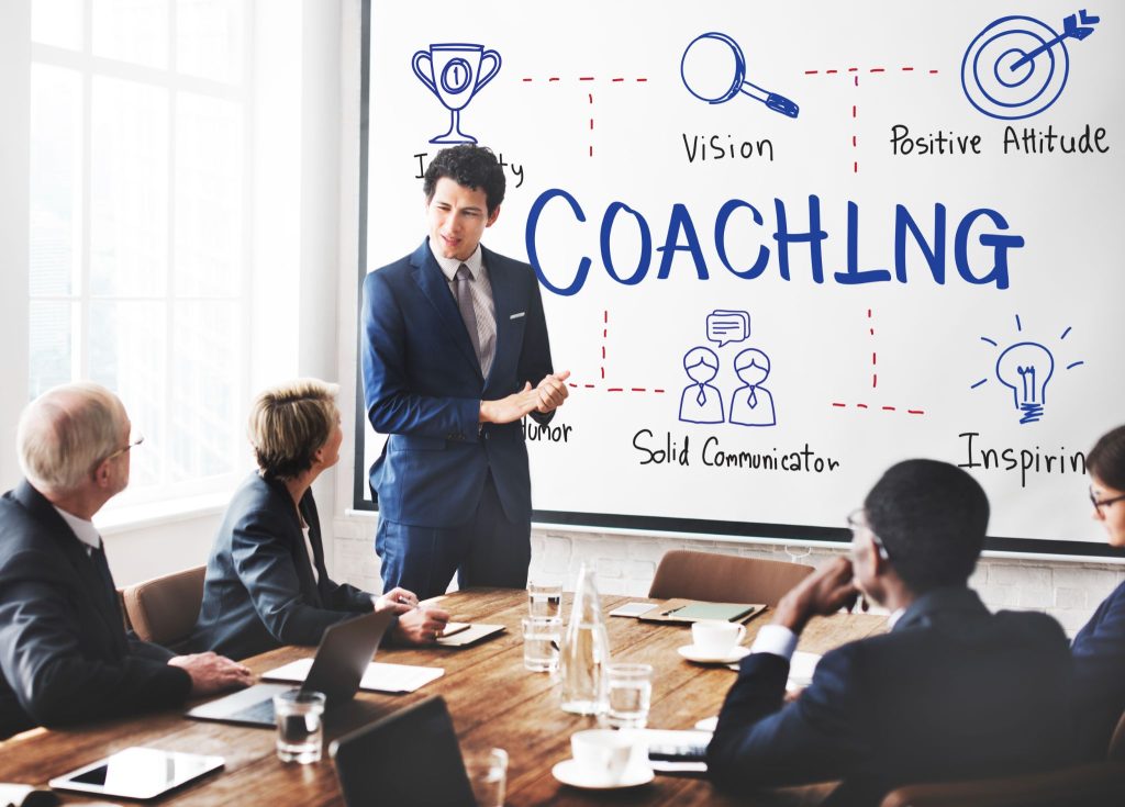 A business professional giving a presentation on coaching to a group, with key points like vision, communication, and positive attitude displayed on a whiteboard.