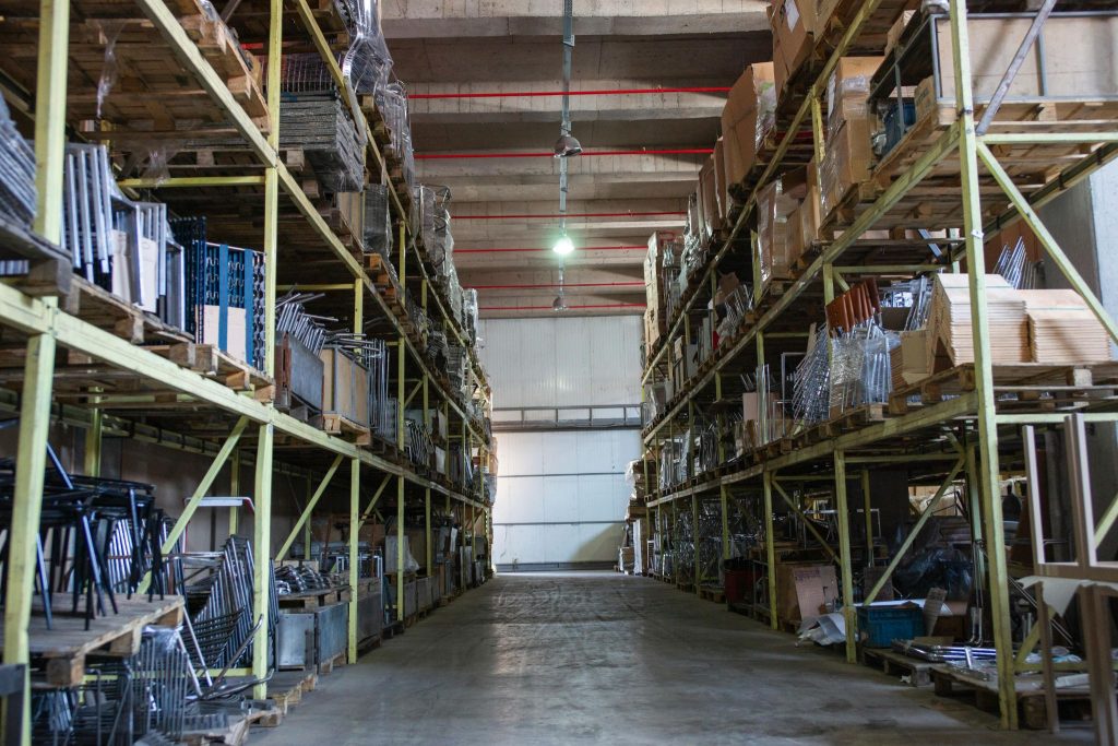 A long aisle in a warehouse with rows of shelves filled with boxes and pallets. The shelves are tall and reach almost to the ceiling. The lighting is dim, and the air is musty.