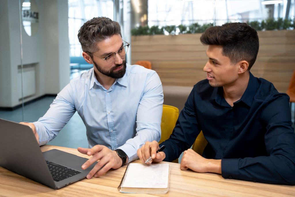 Two colleagues in an office having a focused discussion, one pointing at a laptop screen while the other takes notes.